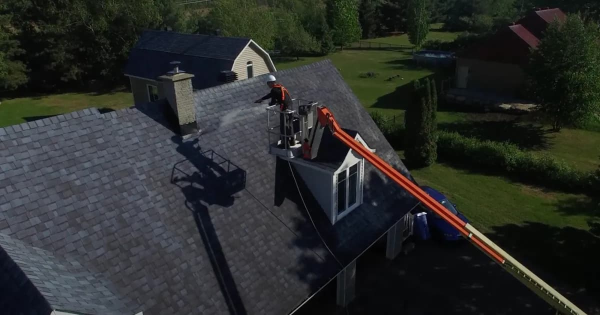 An applicator in a boom lift spraying a treatment onto an asphalt shingle roof, seen from above