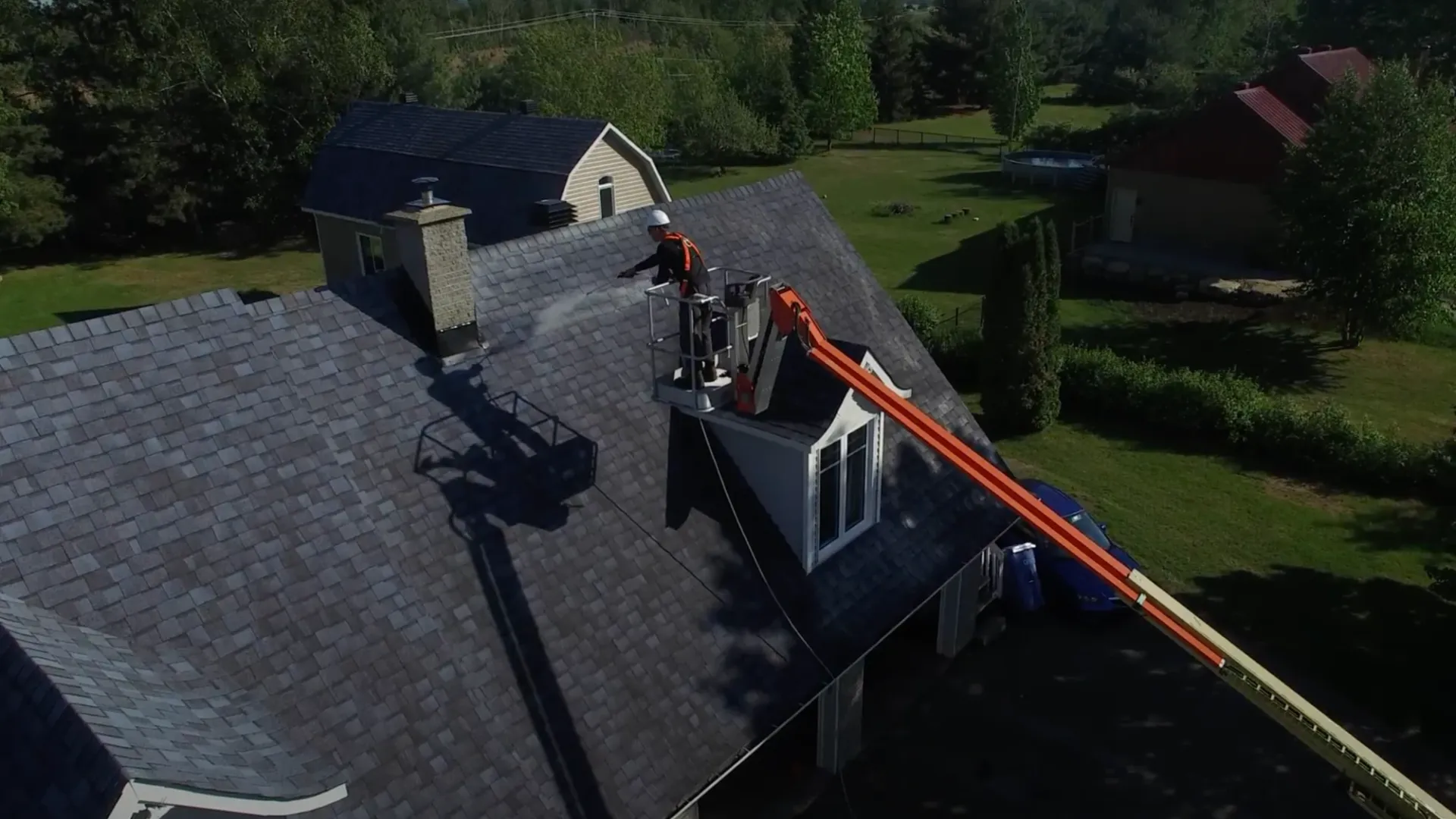 An applicator in a boom lift spraying a treatment onto an asphalt shingle roof, seen from above